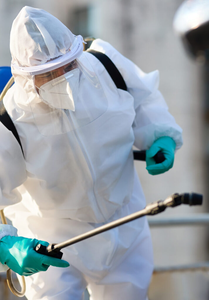 Sanitation worker in hazmat suit spraying car with disinfectant due to COVID-19 epidemic.